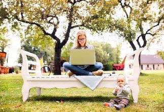 woman sitting on an outside bench with a laptop open and her young child sitting on the grass